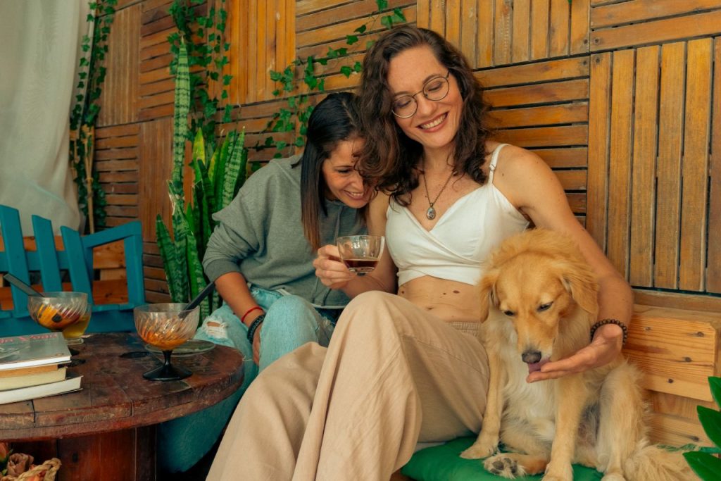 Welcome to the Society of Friends Two women enjoying drinks and laughter with a dog in a cozy indoor setting.